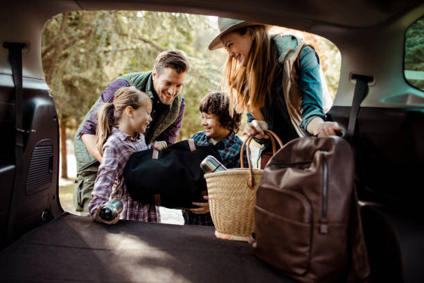 A cheerful family loading items into the back of their car for a trip. The parents and two children are smiling, dressed casually for outdoor activities, and surrounded by camping gear, showing excitement for an upcoming adventure.