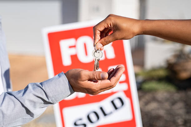 Close-up of two people exchanging keys, one hand passing the house keys to another with a 'For Sale' sign that reads 'Sold' in the background.