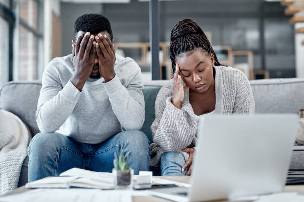 A distressed couple sitting on a couch, looking at documents and a laptop, with their heads in their hands, indicating frustration or concern, possibly over financial issues or home-related matters.