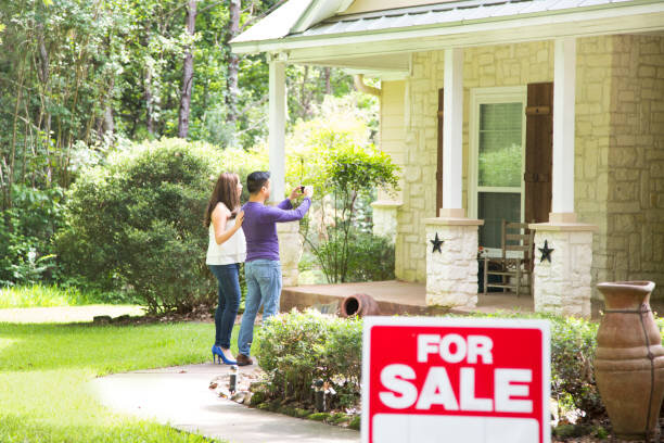 A young couple standing outside a house that is for sale, taking pictures and inspecting the property. A prominent "For Sale" sign is visible in the foreground. The house features a spacious front porch and a manicured lawn, surrounded by lush greenery.