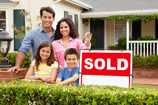 A happy family of four standing in front of their new home, smiling as they proudly hold a key and pose next to a large red 'SOLD' sign. The children are beaming with joy, symbolizing the excitement of a successful home purchase.