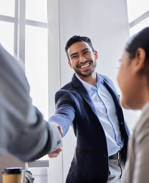 A confident, smiling businessman in a navy-blue suit shakes hands with another individual in a modern office setting, conveying a successful agreement or positive business interaction. The man’s expression radiates friendliness and professionalism, enhancing the welcoming and engaging atmosphere of the scene.