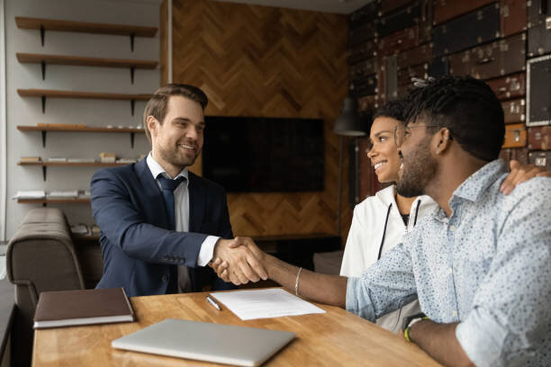 A real estate agent in a dark blue suit is shaking hands with a smiling couple seated across a wooden table in a modern office. The couple, appearing happy and satisfied, is leaning forward, with the woman gently resting her hand on the man’s shoulder. A few documents are laid out on the table, symbolizing the conclusion of a successful real estate transaction.