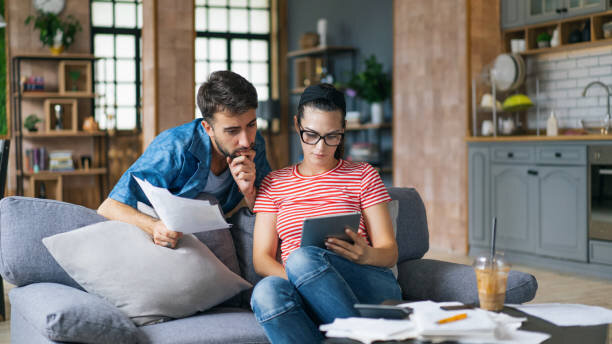 Young couple sitting on a couch in a modern living room, reviewing financial documents and using a tablet, appearing focused and concerned.