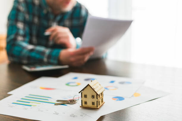 A small wooden house model with a set of keys on top, placed on a desk with charts and graphs scattered around. In the blurred background, a man in a plaid shirt reviews financial documents, indicating a focus on real estate investments or property analysis.