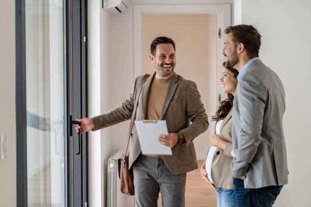 A smiling real estate agent welcoming a couple into a new home. The agent holds a clipboard and gestures towards the entrance, while the couple looks excited and engaged. The couple, dressed in business casual attire, stands close together, highlighting their interest in the property. The agent's positive demeanor and professional appearance create a welcoming atmosphere, indicating a successful showing or property tour.