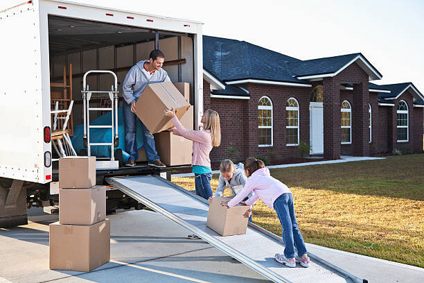 A family loading boxes onto a moving truck. The parents and children are working together, moving into a new home with a modern brick exterior.