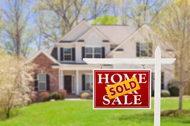 A red and white "Home for Sale" sign with a "SOLD" sticker in front of a beautiful suburban house, indicating that the property has been successfully sold.