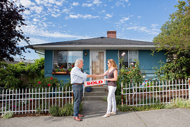 An elderly man and a woman shaking hands in front of a charming blue house with a white picket fence and garden flowers, celebrating the sale of the property. The woman is holding a red "SOLD" sign, indicating a successful home transaction under a clear sky.