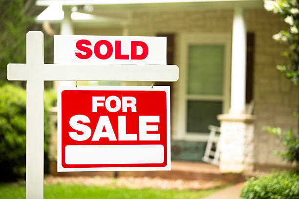 A close-up of a "Sold" sign over a "For Sale" sign, standing in front of a home. The background is slightly blurred, with a glimpse of a porch and rocking chair visible.