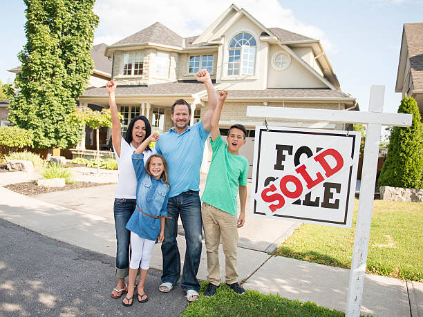 A happy family of four, standing in front of a house they just purchased. The family is smiling and celebrating their new home ownership, with the father and mother raising their hands in excitement. A large "SOLD" sign is visible in the foreground, signifying the successful sale of the property.