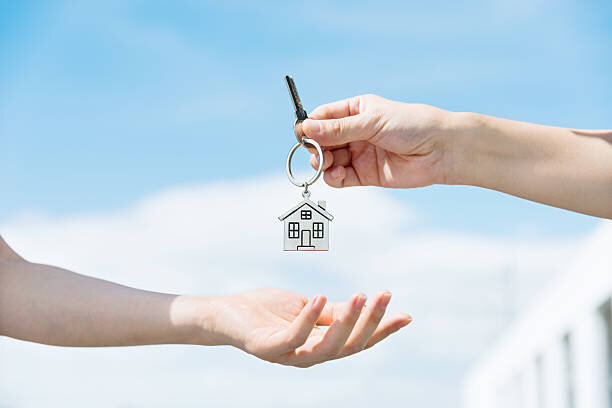 Close-up of a hand passing over a house-shaped keychain with a key to another hand, symbolizing the transfer of property ownership or renting a new home.
