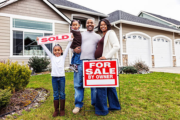 A happy family of four standing in front of a newly sold house, holding a "SOLD" sign. The father, holding a young child, smiles alongside the mother and an excited daughter who is raising the sign with joy. A "For Sale By Owner" sign is positioned in front of the well-kept lawn, and the modern house exterior features large windows and a garage.
