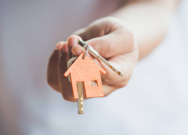 A hand holding a house-shaped keychain with a set of keys, symbolizing homeownership or the transfer of property keys after a sale.