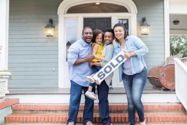 Happy family of four standing on the front porch of their newly purchased home, holding a 'SOLD' sign. The family includes a mother, father, and two young children, all smiling and celebrating their recent home purchase. The house in the background features a modern design with green siding and white trim, giving a welcoming feel.