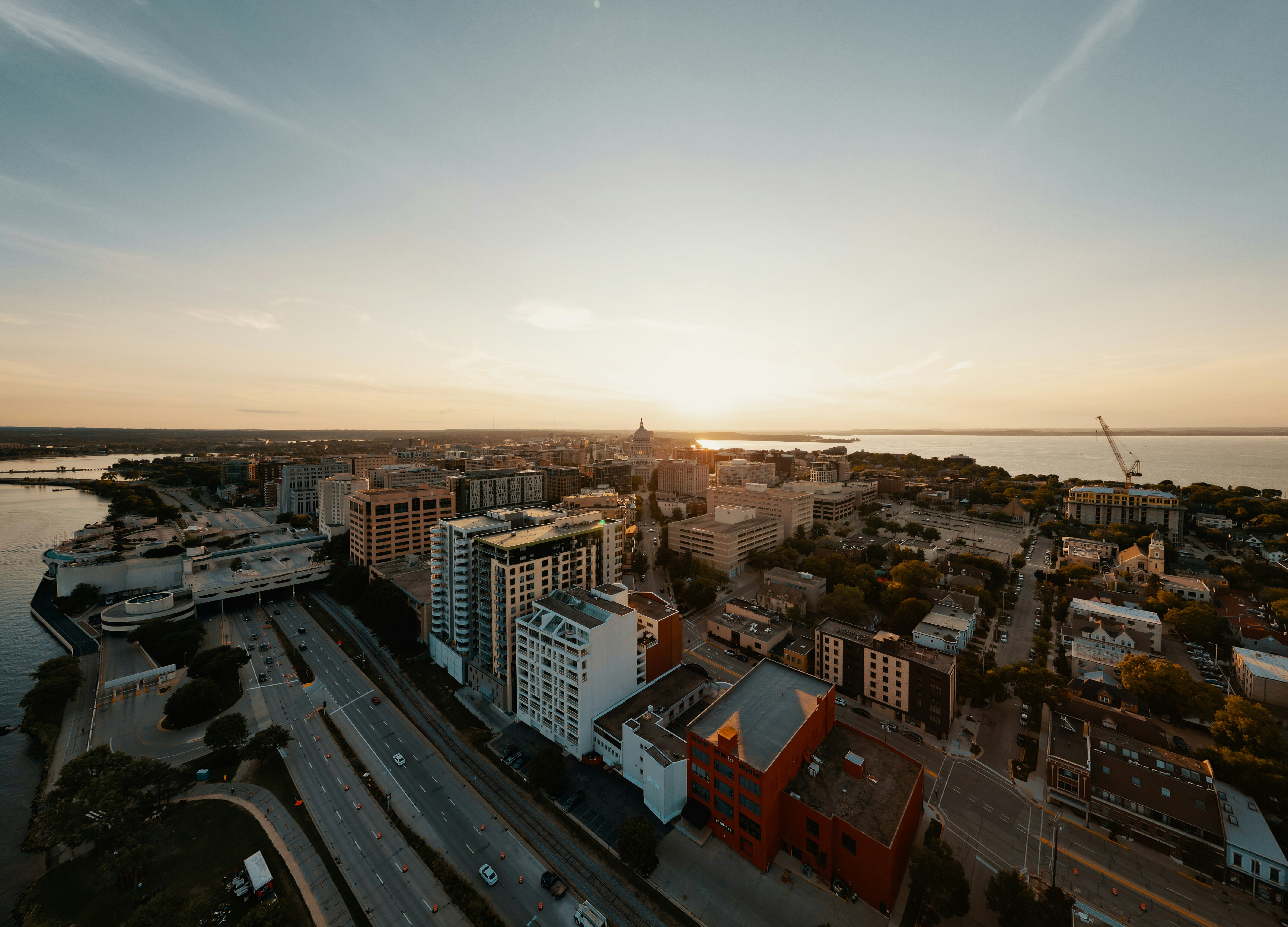 A panoramic aerial view of a cityscape at sunset, featuring downtown buildings, roads, and a body of water in the distance, evoking an urban landscape scene at dusk.