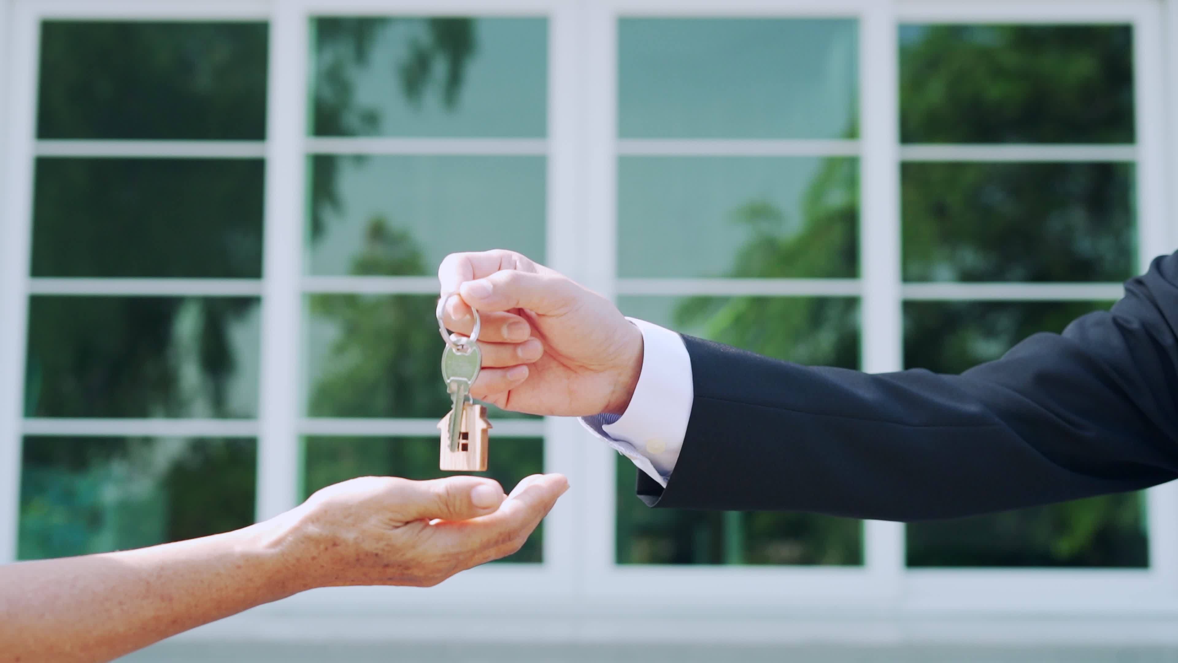 A close-up shot of a professional real estate agent handing over a house key with a wooden home-shaped keychain to a new homeowner. The agent is wearing a suit, and the background features a modern house facade with large glass windows, symbolizing the transfer of property ownership.
