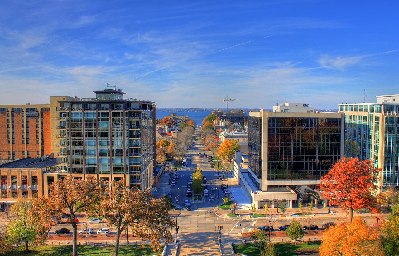  An elevated view of downtown Madison, Wisconsin, featuring tall buildings, colorful trees, and a distant view of Lake Monona under a clear blue sky. The bright autumn colors enhance the picturesque urban landscape.