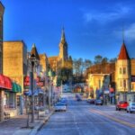 Vibrant downtown scene of a small town with colorful buildings, shops, and a church steeple in the background, set under a clear blue sky. Cars are parked along the street, and small businesses line the sidewalk.