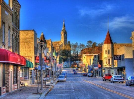 Vibrant downtown scene of a small town with colorful buildings, shops, and a church steeple in the background, set under a clear blue sky. Cars are parked along the street, and small businesses line the sidewalk.