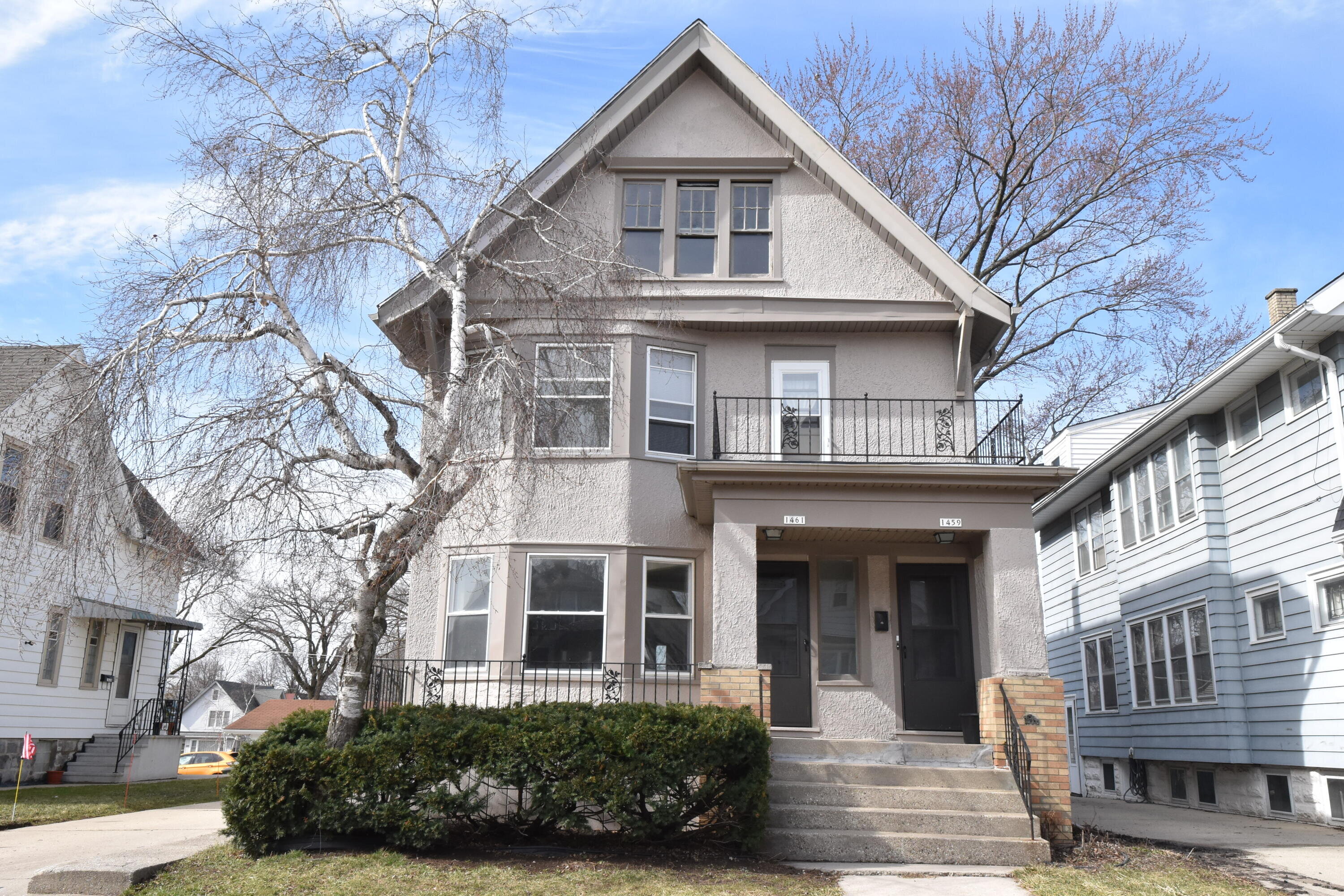 A two-story duplex-style home with beige stucco walls and a classic architectural style. A mature birch tree with branches reaching toward the house frames the front view. A small balcony and steps lead up to the entrance, with lush green bushes bordering the front yard.