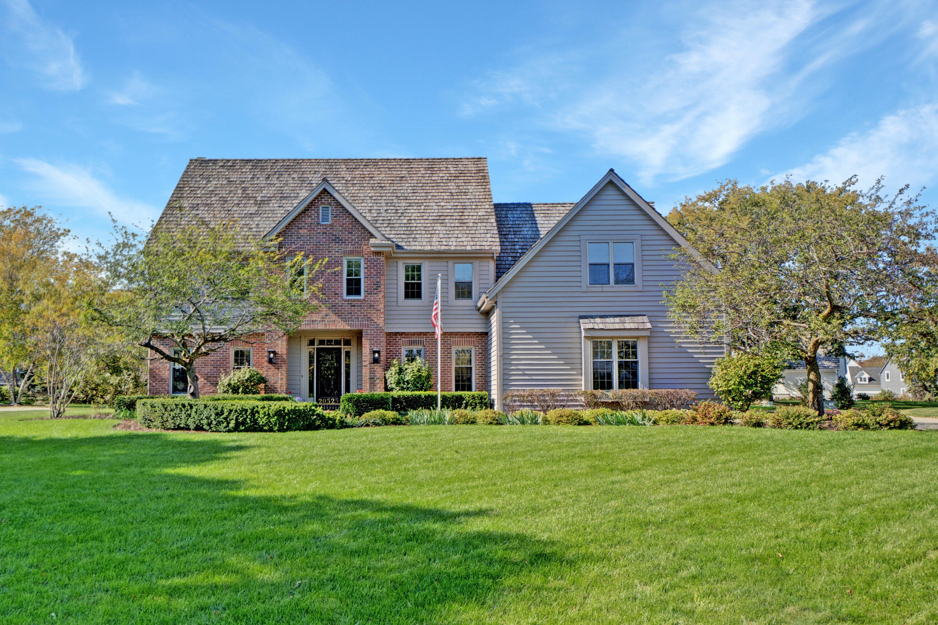 A spacious two-story suburban home with a blend of brick and siding exterior. The house has a high-pitched roof with wooden shingles, large windows, and a manicured front lawn. The entrance is framed by shrubs and a flagpole, giving the property a welcoming and patriotic feel.
