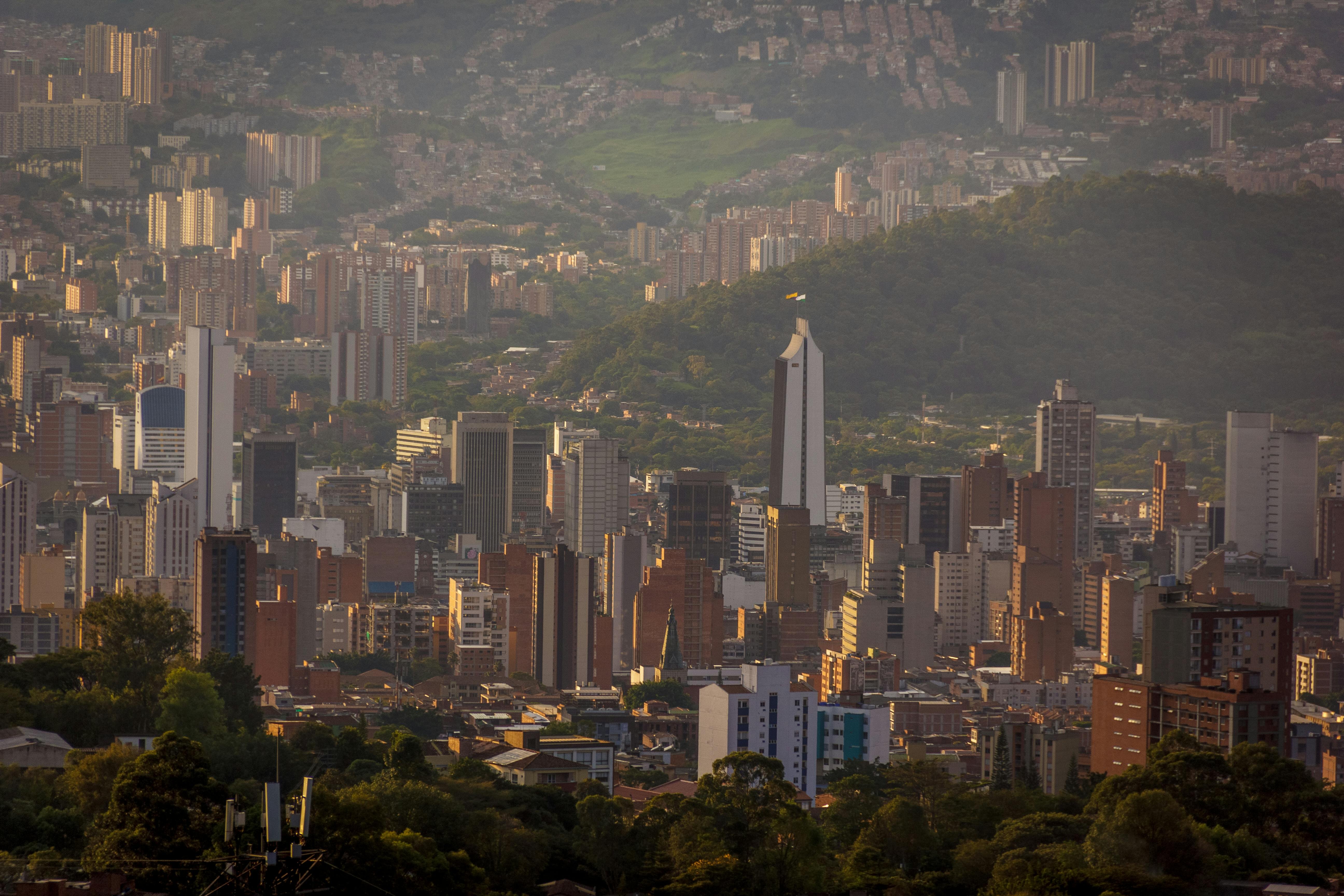 A distant view of a densely populated city skyline against a lush green mountainous backdrop, showcasing a blend of tall residential and commercial buildings under sunlight, creating a dramatic contrast between urban and natural elements.