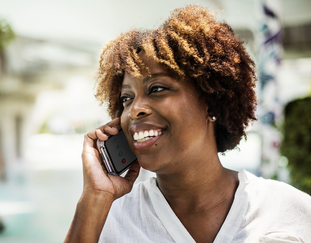 Close-up of a cheerful woman with short curly hair, smiling while holding a smartphone to her ear as she makes a call. The background is softly blurred, highlighting the woman's positive expression, suggesting a friendly conversation or good news.