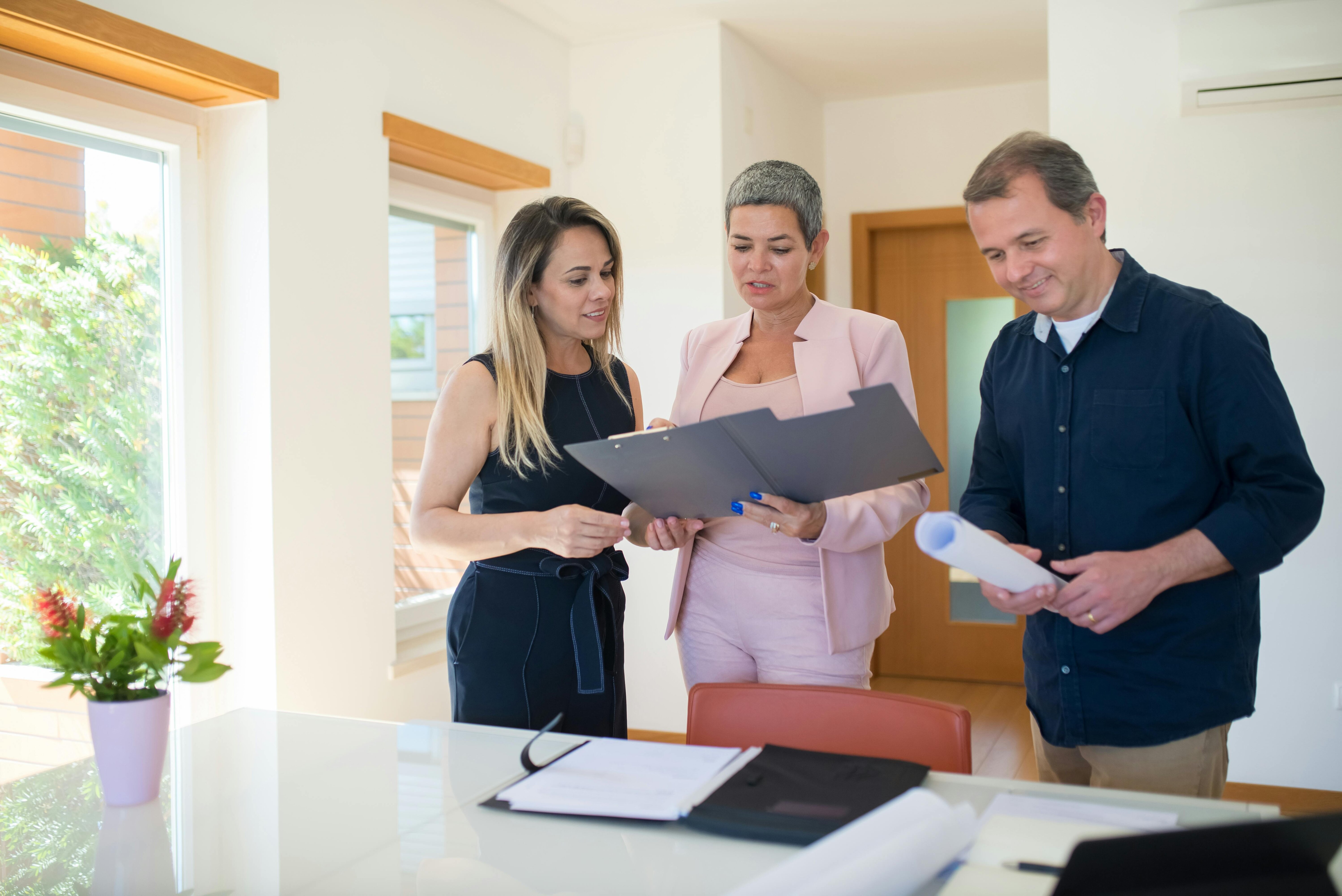 Three business professionals discussing documents inside a modern office. A woman in a pink suit is holding a folder and reviewing papers with her colleagues, while another woman in a black dress and a man in casual attire listen attentively. The setting includes natural light and a view of greenery through large windows.