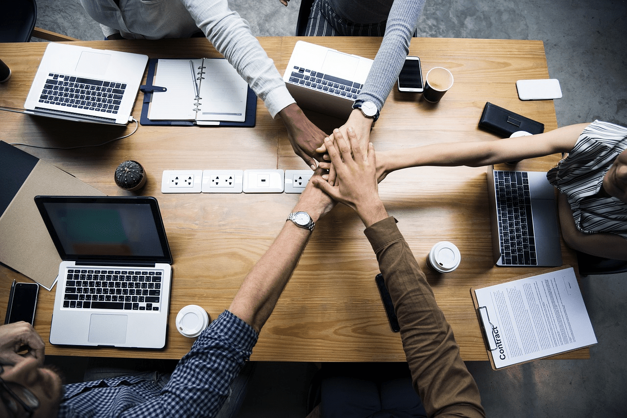 A group of business professionals sitting around a large wooden table, stacking their hands together in a display of teamwork and collaboration. The table is covered with laptops, notebooks, and coffee cups.