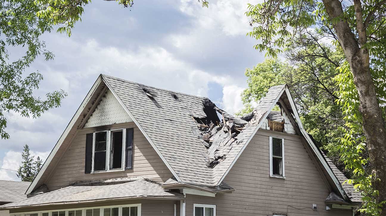 A house with severe roof damage, showing a large hole and missing shingles, possibly due to a natural disaster or fire damage, with trees in the background.