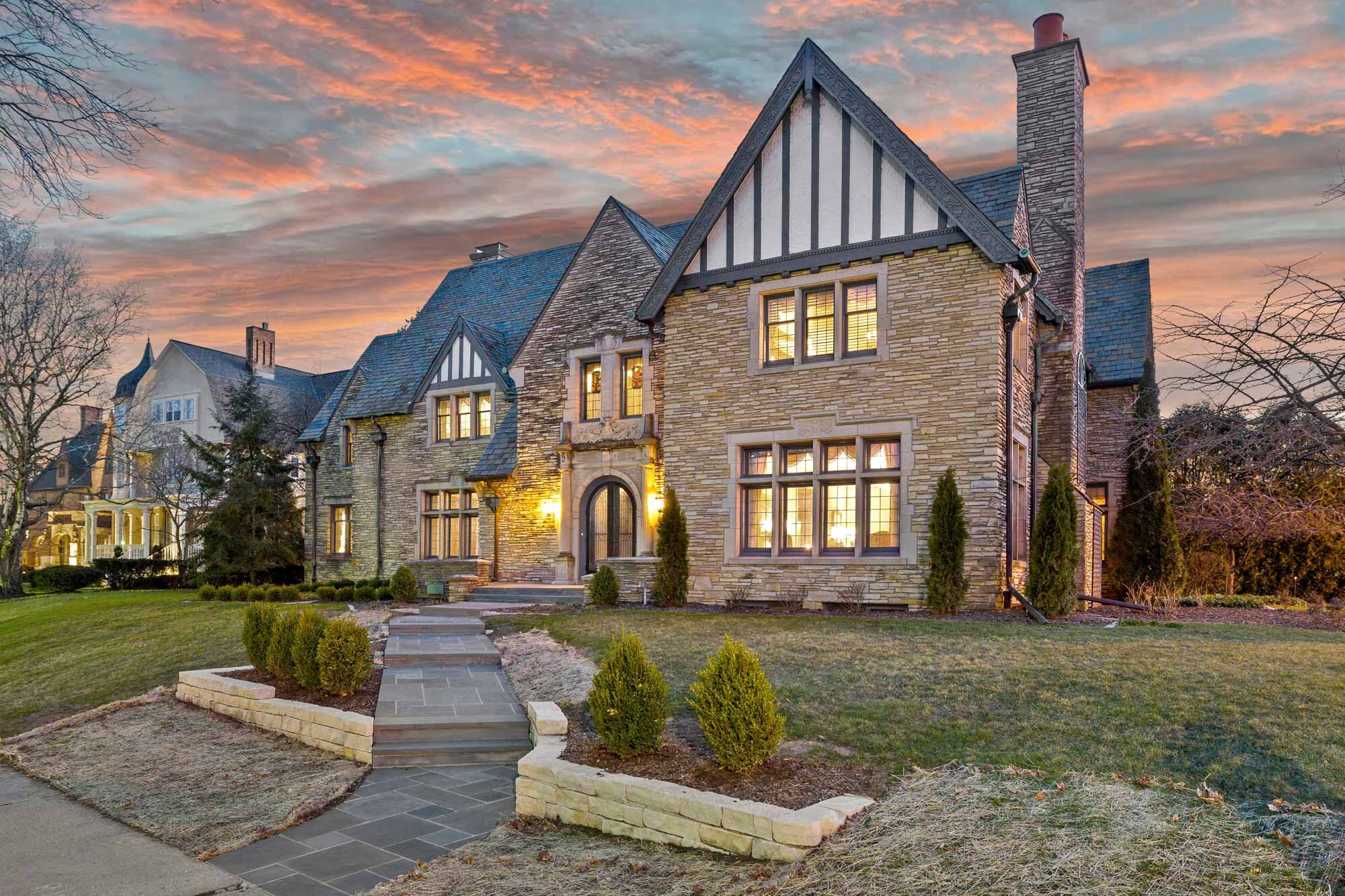 A grand Tudor-style house with a stone facade, prominent gabled rooflines, and intricate wood detailing, captured during sunset with a pink and orange sky. The well-maintained front yard features a pathway, small manicured bushes, and mature trees.