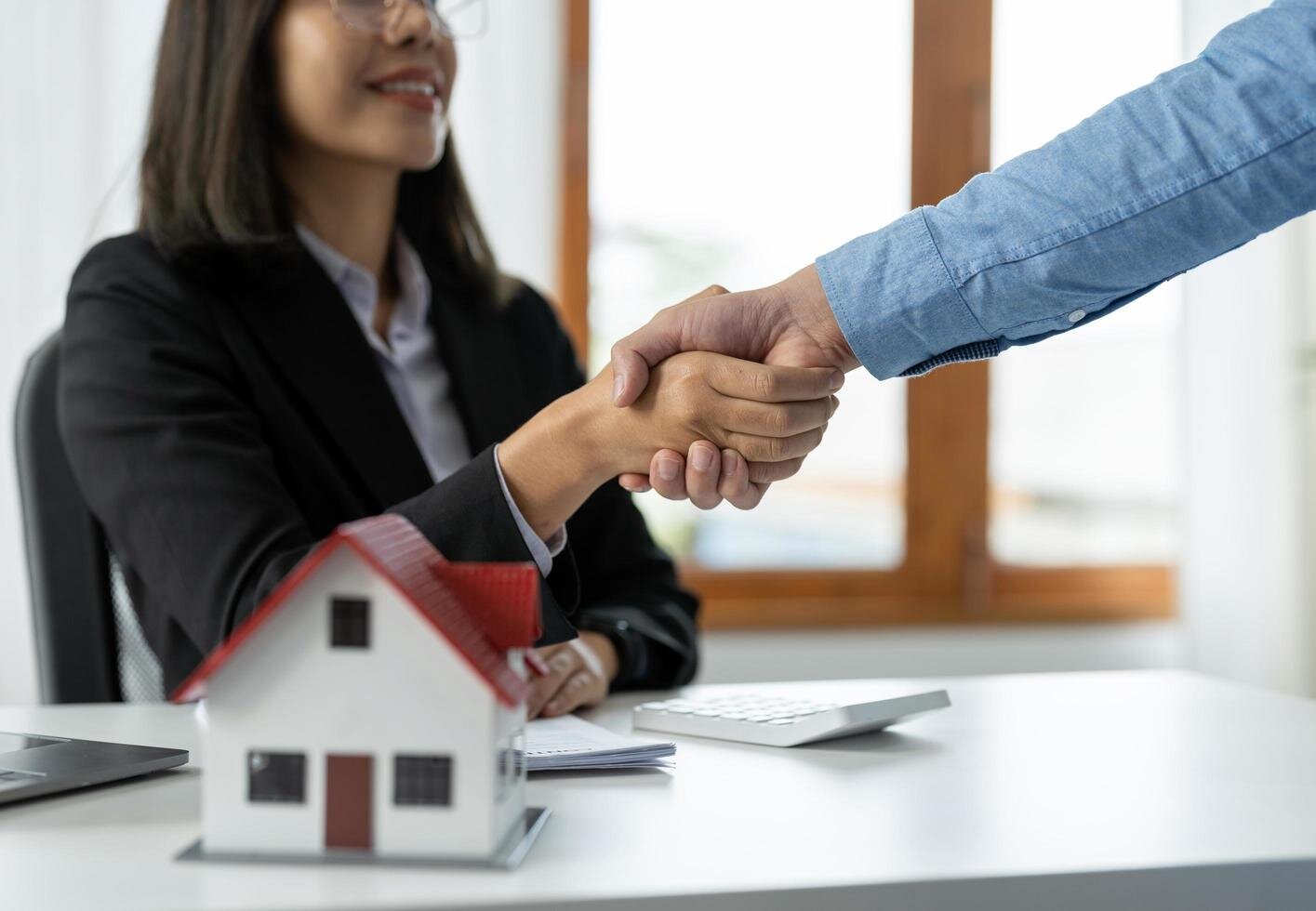 A real estate agent and a client shaking hands over a small house model on a desk, symbolizing a successful property deal or agreement.