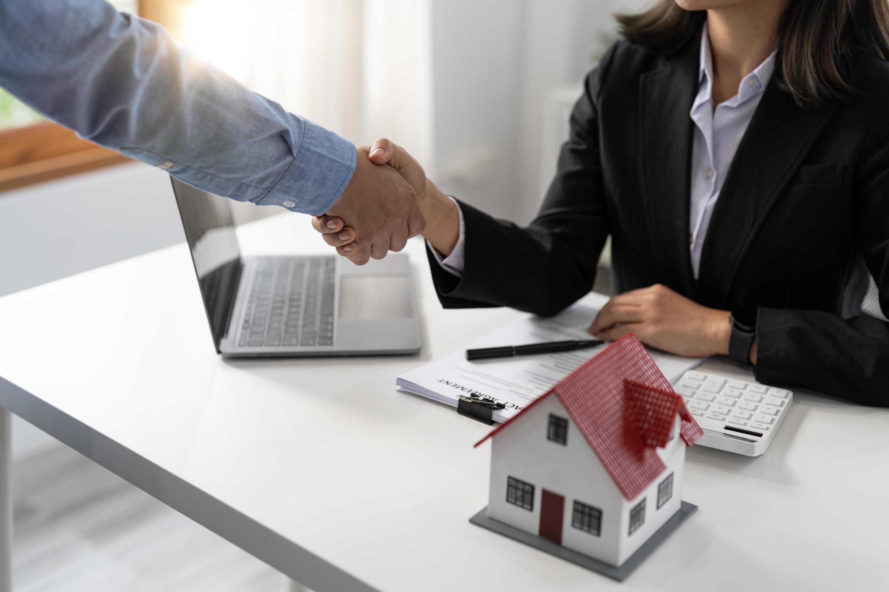A close-up shot of a handshake between a man in a blue shirt and a businesswoman dressed in a black suit in a modern office setting. A small model house with a red roof and a laptop are visible on the desk, suggesting a real estate transaction.