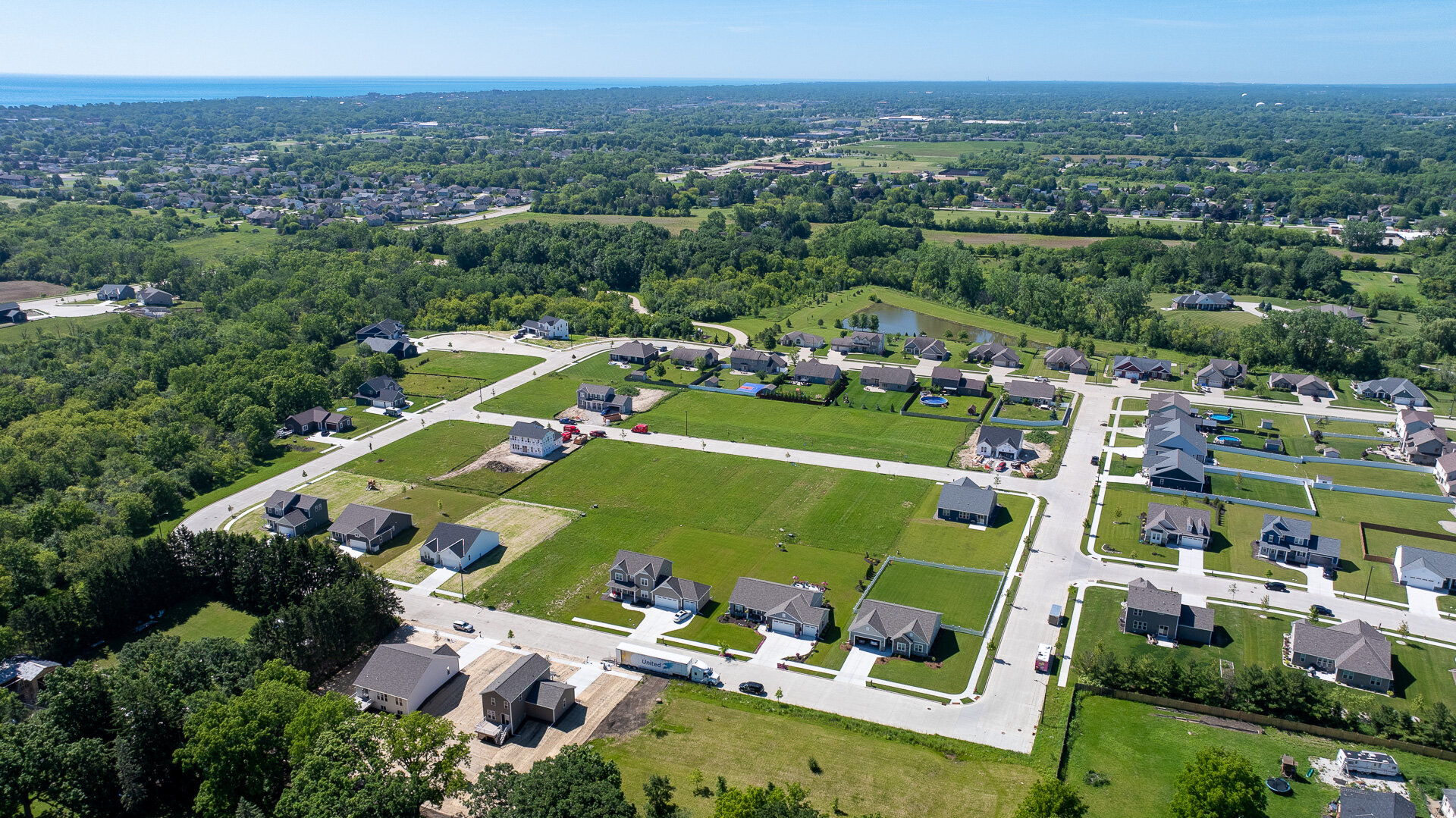 A bird's-eye view of a newly developed residential neighborhood with spacious homes on large lots, tree-lined streets, and nearby greenery. The community is nestled in a scenic landscape with a distant view of a body of water on the horizon.