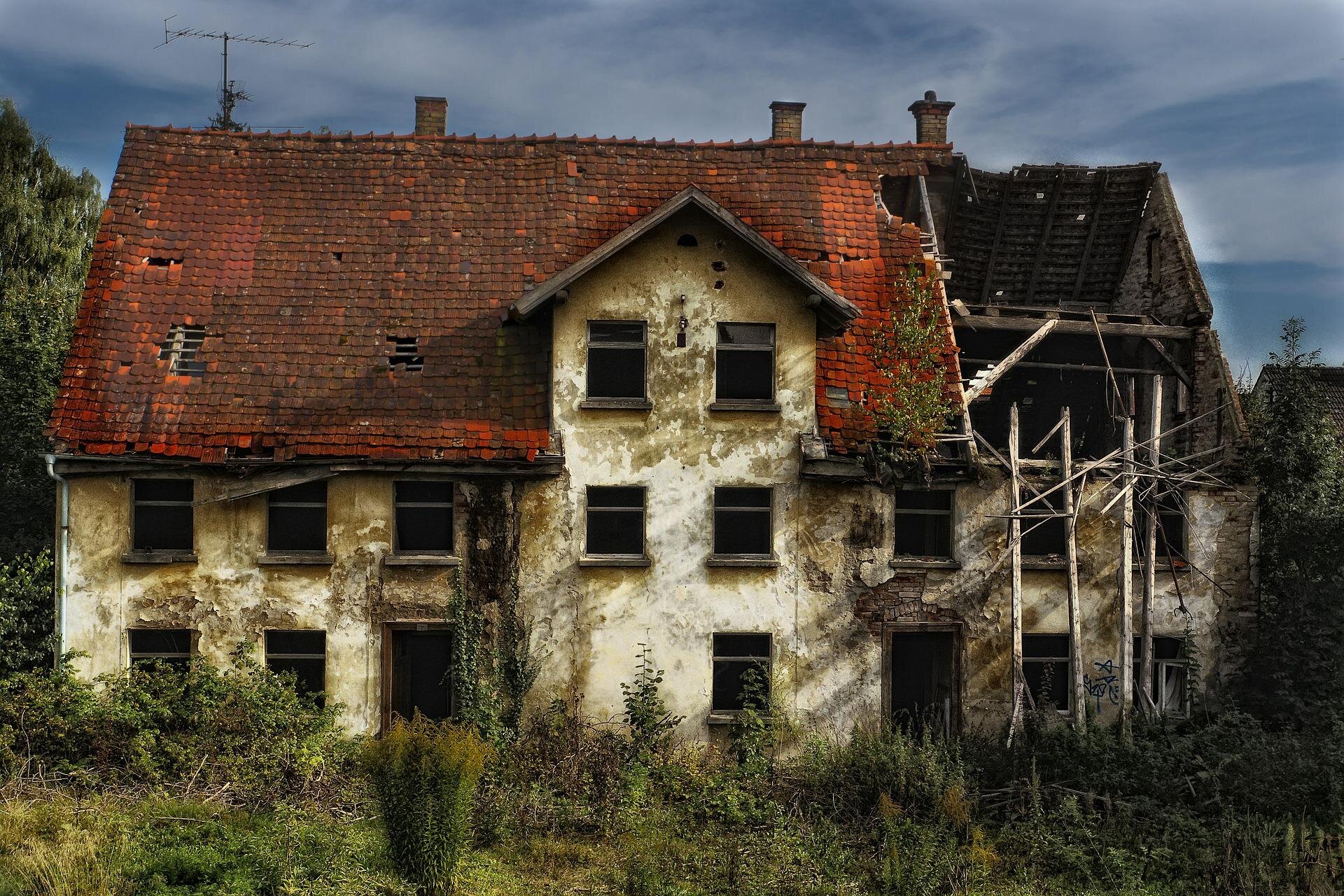 A dilapidated house with a severely damaged roof and overgrown vegetation surrounding it, showcasing signs of abandonment and decay.