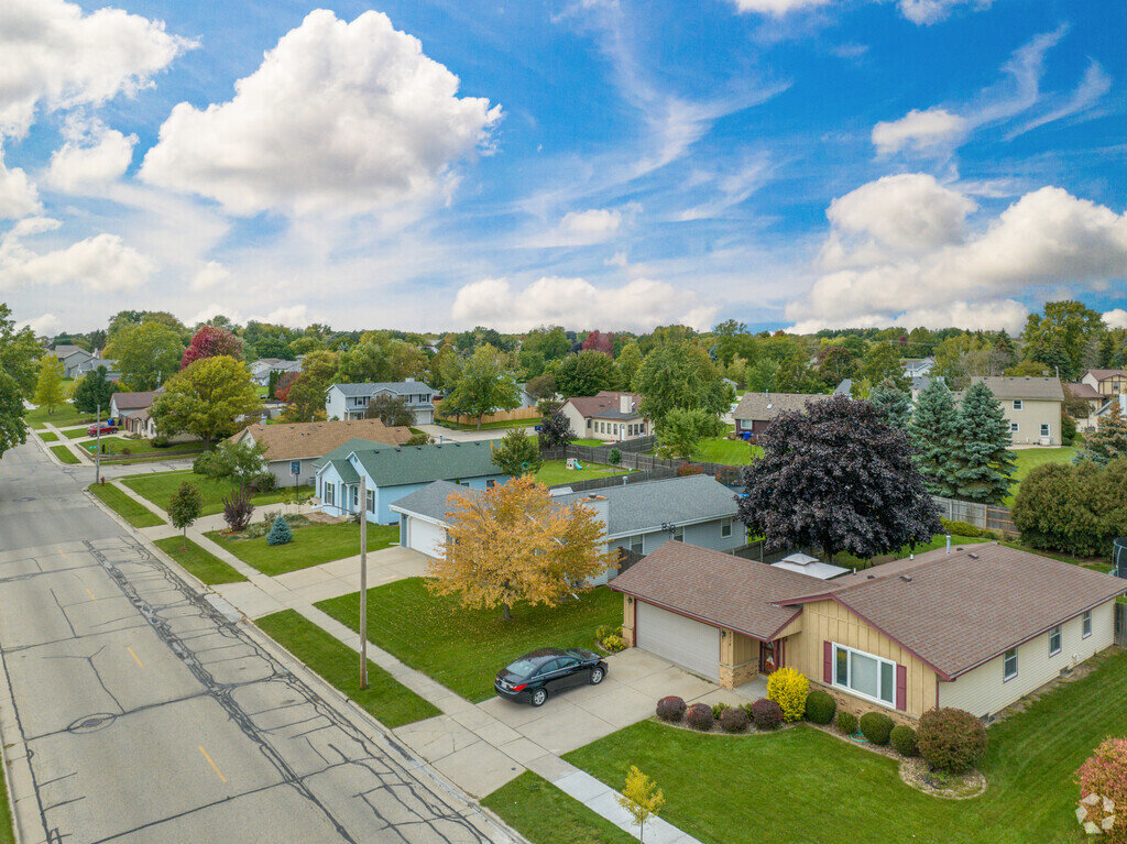Aerial view of a quiet suburban neighborhood with single-family homes lined along a tree-shaded street. The houses are well-kept, and the lawns are neatly manicured, reflecting a serene and well-maintained community under a partly cloudy blue sky.