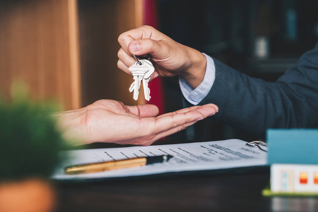A close-up image of a professional handing over a set of house keys to another person. The recipient's hand is open, palm facing up, while the giver holds the keys delicately. The background includes a table with paperwork, symbolizing the completion of a real estate transaction.