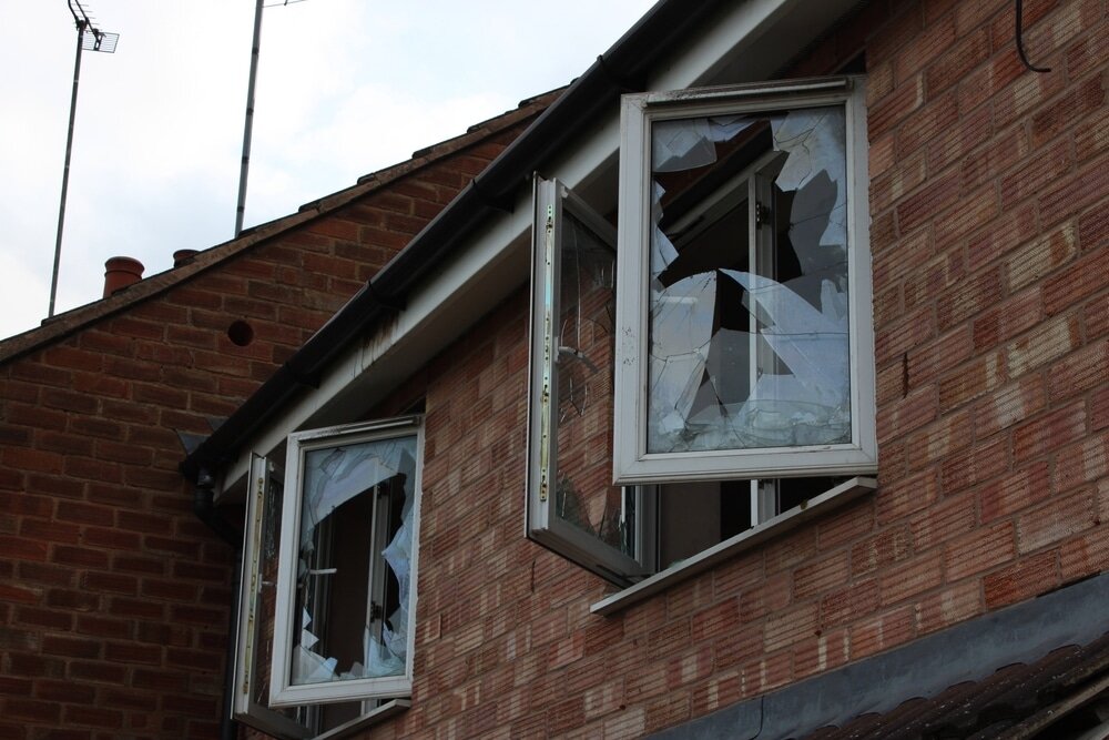 A two-story brick house with broken windows and damaged frames. The open windows reveal shattered glass panels, indicating a state of disrepair or vandalism. The image highlights property damage, making the house appear uninhabitable.
