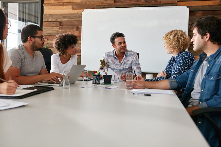 A group of six people sitting around a table in a modern office setting, engaged in a collaborative discussion. The whiteboard behind them suggests a brainstorming or team meeting environment. The casual atmosphere reflects a creative work setting.