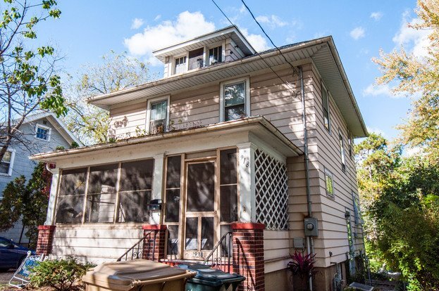 A two-story beige house with a screened-in front porch and red brick pillars at the entrance. The house has a classic design with multiple windows and a slightly worn exterior. Tall trees and some green plants surround the home.