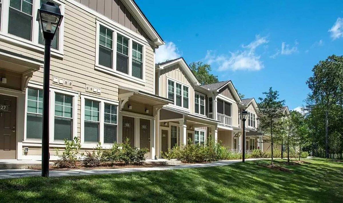 A row of modern, well-maintained townhouses with beige and gray exteriors. The houses are surrounded by manicured greenery and a bright blue sky, creating a welcoming neighborhood feel. Each home has individual porches and windows with white trim, emphasizing a sense of uniformity and community.