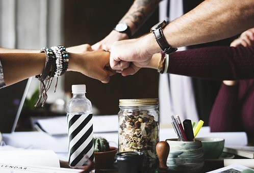A close-up view of multiple hands coming together for a fist bump over a wooden office table. The workspace is filled with office supplies, including notebooks, pens, a glass jar, a water bottle, and a potted cactus, symbolizing teamwork and collaboration.