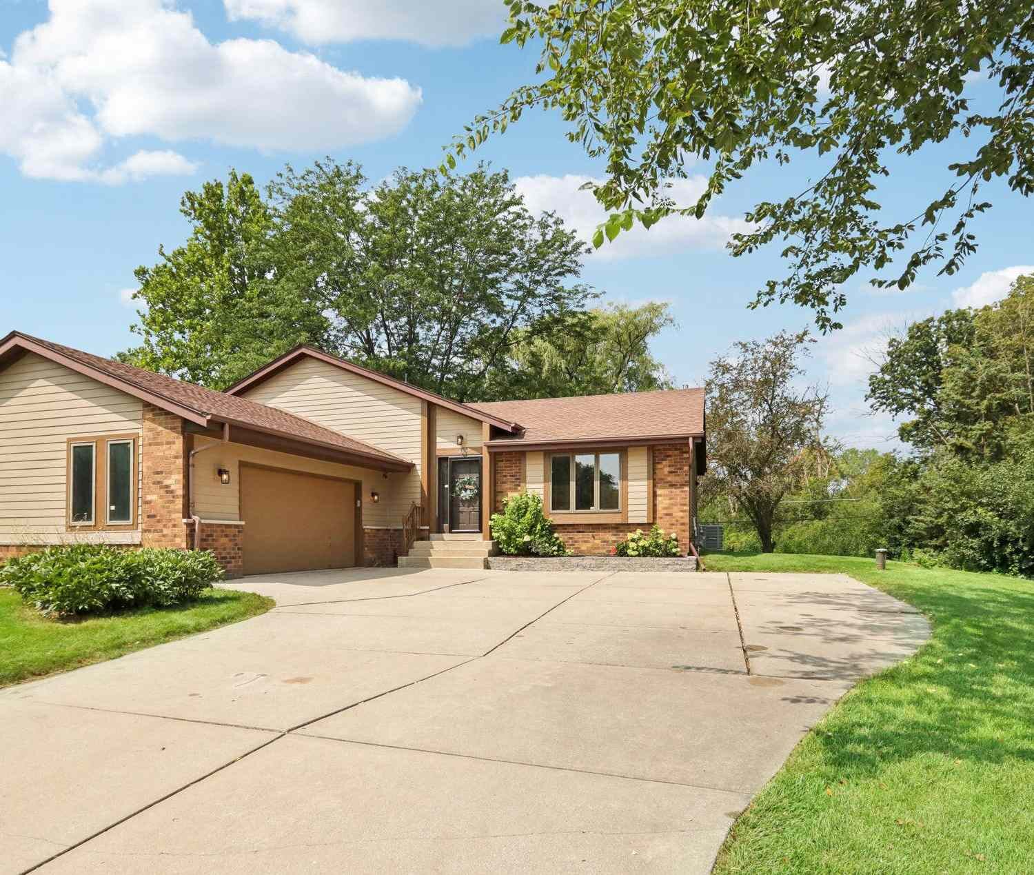 A single-story suburban home with a light beige exterior and a two-car garage. The house features a sloped roof and is set back from the road with a wide driveway and a manicured lawn surrounded by trees.
