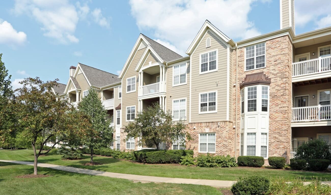 Modern apartment complex with beige siding, multiple balconies, and landscaped greenery in front, under a partly cloudy sky.