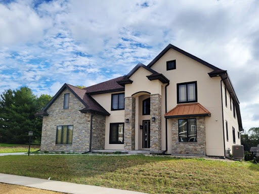 A modern two-story house with a combination of stone and beige stucco exterior. The house has a unique architectural design with a prominent entryway, black window frames, and a blend of modern and rustic elements. The surrounding lawn is green with a paved walkway leading to the front door.