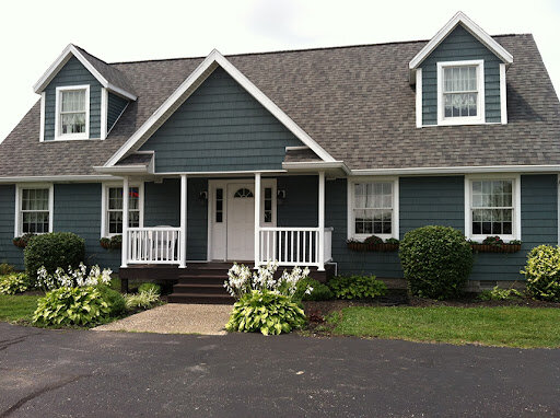 A cozy Cape Cod-style house with green siding and white trim, featuring a welcoming front porch and two dormer windows on the roof. The landscape includes neatly trimmed shrubs and white flowering plants along the front, with a paved driveway leading to the house.