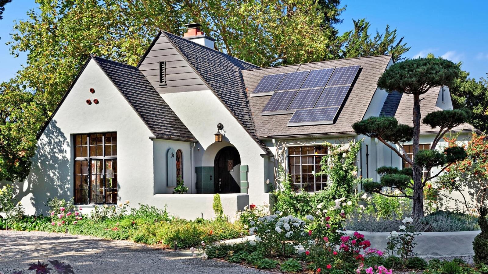 A picturesque, storybook-style cottage with cream stucco walls and a slate roof, adorned with solar panels. The front yard is lush with greenery, including flowering bushes and well-pruned trees, and the arched front door adds to the quaint and eco-friendly aesthetic of the home.