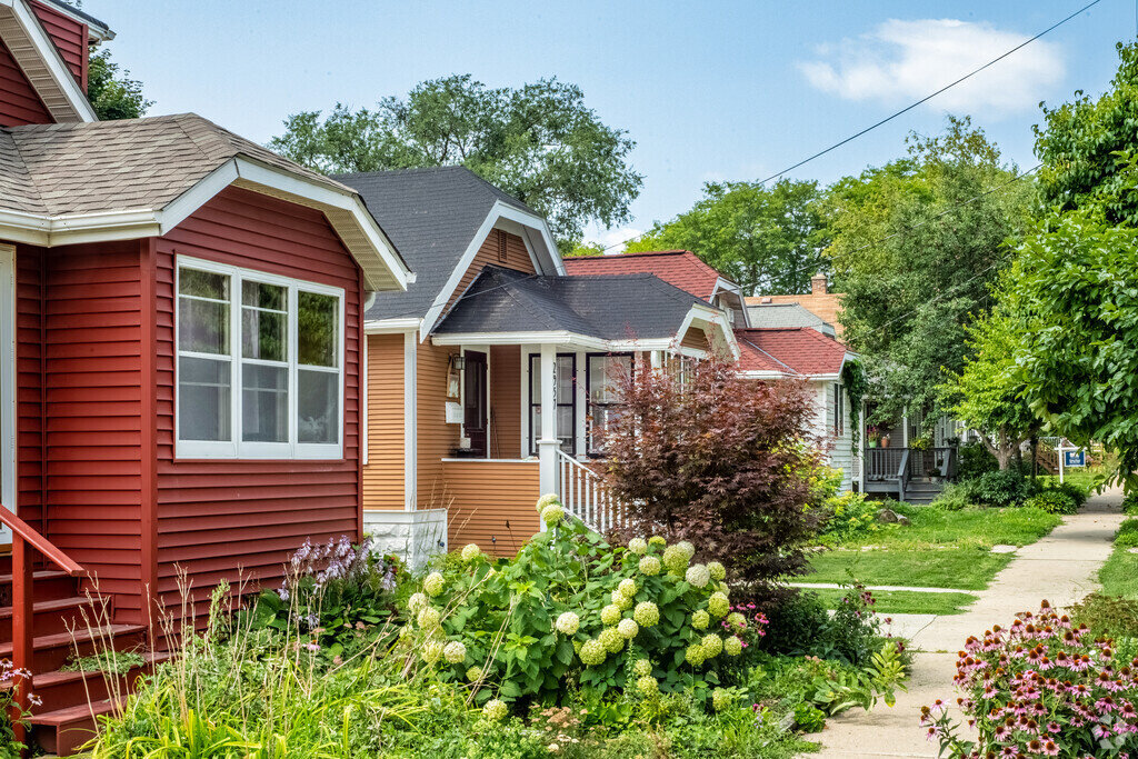 A picturesque row of colorful suburban homes, featuring red, orange, and tan houses with well-maintained front yards and blooming flowers lining the sidewalk.