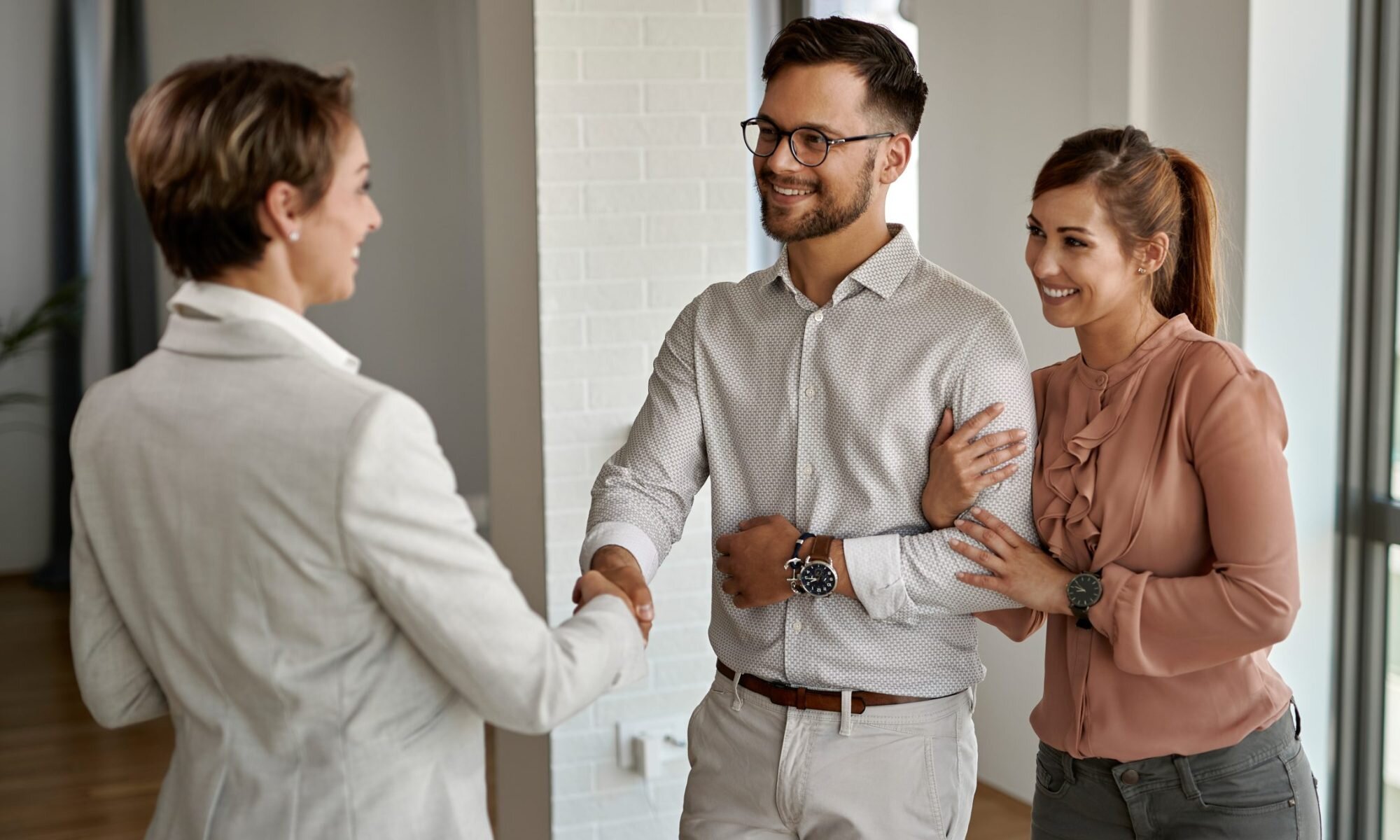A professional real estate agent in a white suit greeting and shaking hands with a young couple inside a modern apartment. The couple, dressed in casual attire, smile warmly, expressing excitement and trust.